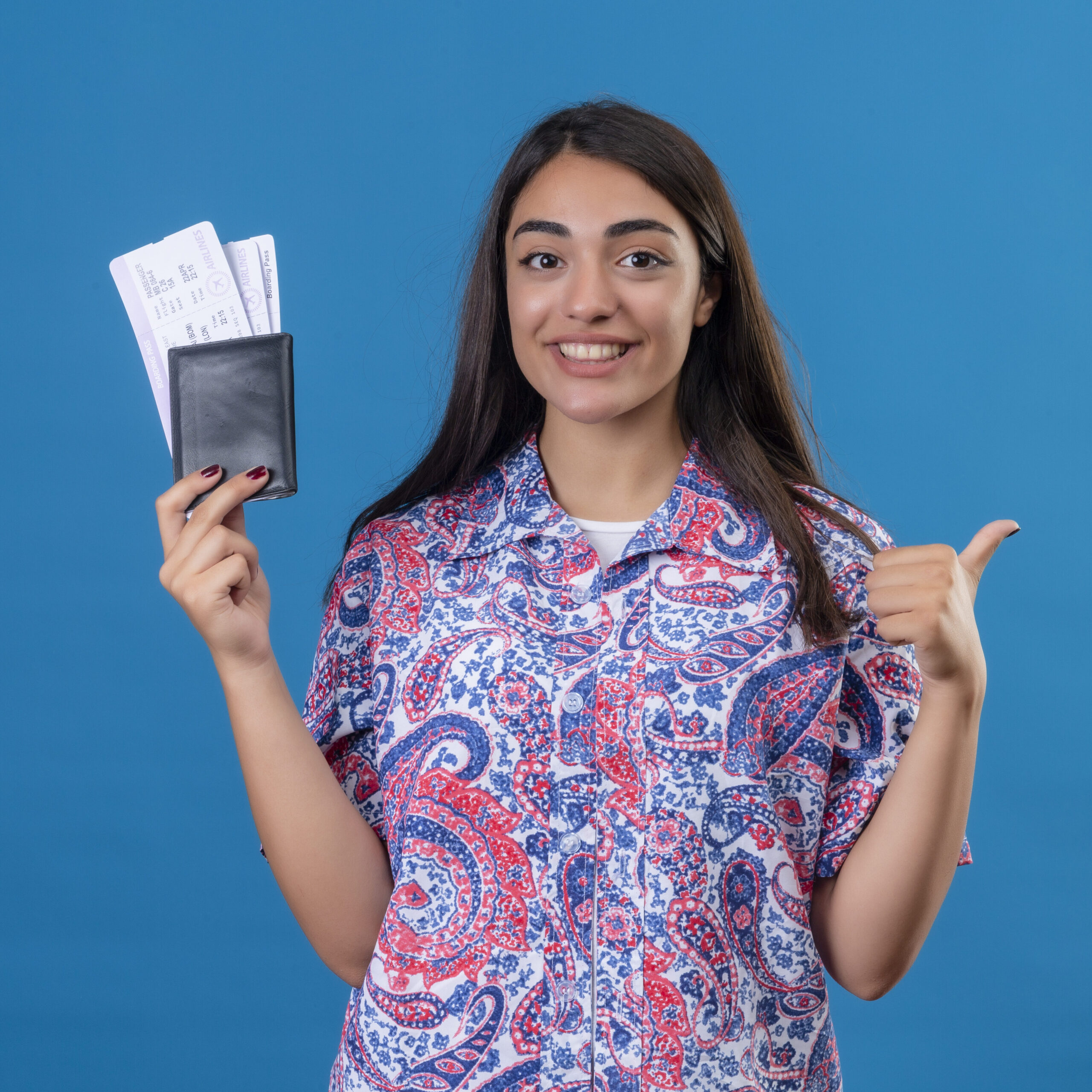 young beautiful woman tourist holding passport with tickets looking at camera smiling cheerfully, showing thumbs up, ready to holiday standing over isolated blue background