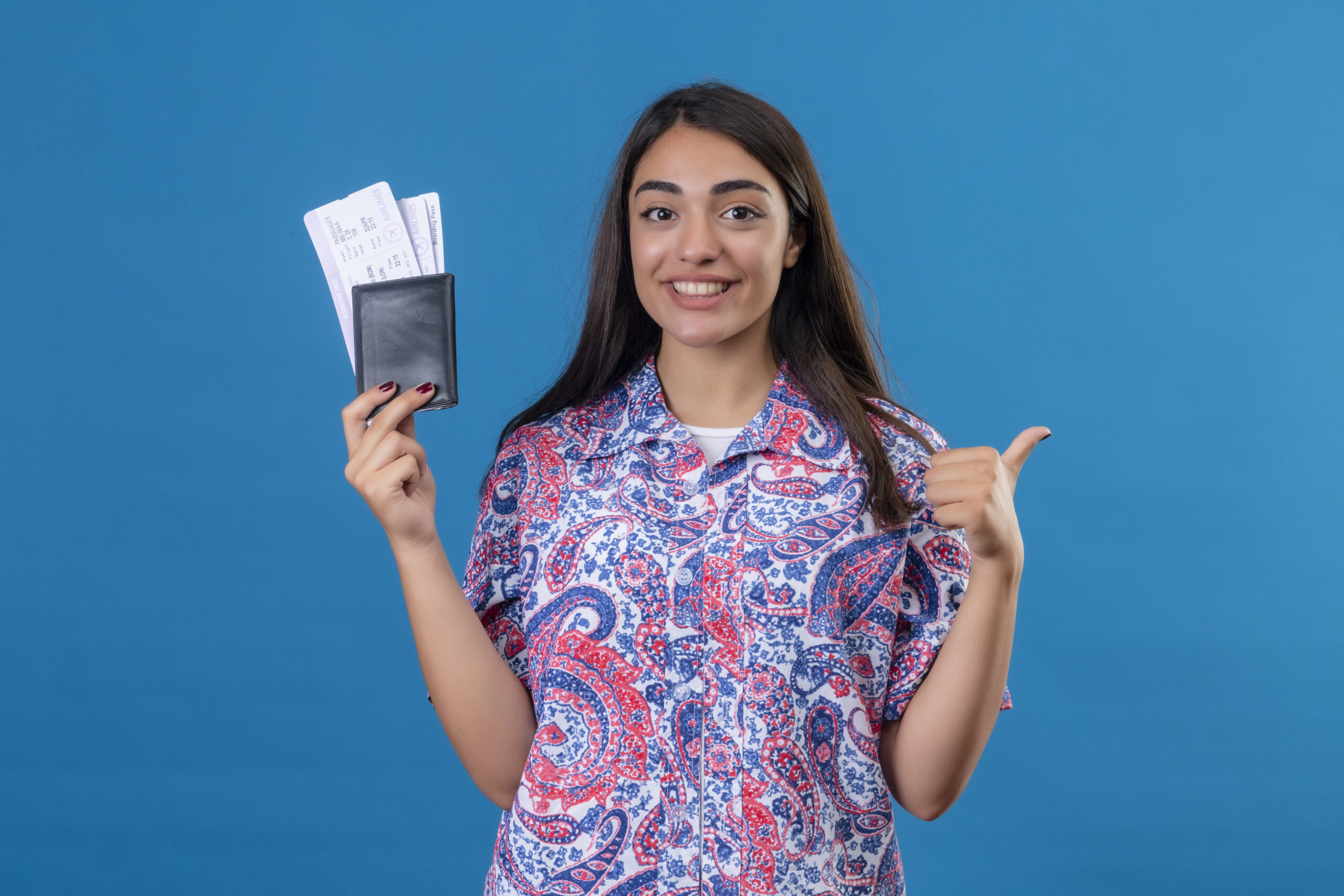 young beautiful woman tourist holding passport with tickets looking at camera smiling cheerfully, showing thumbs up, ready to holiday standing over isolated blue background